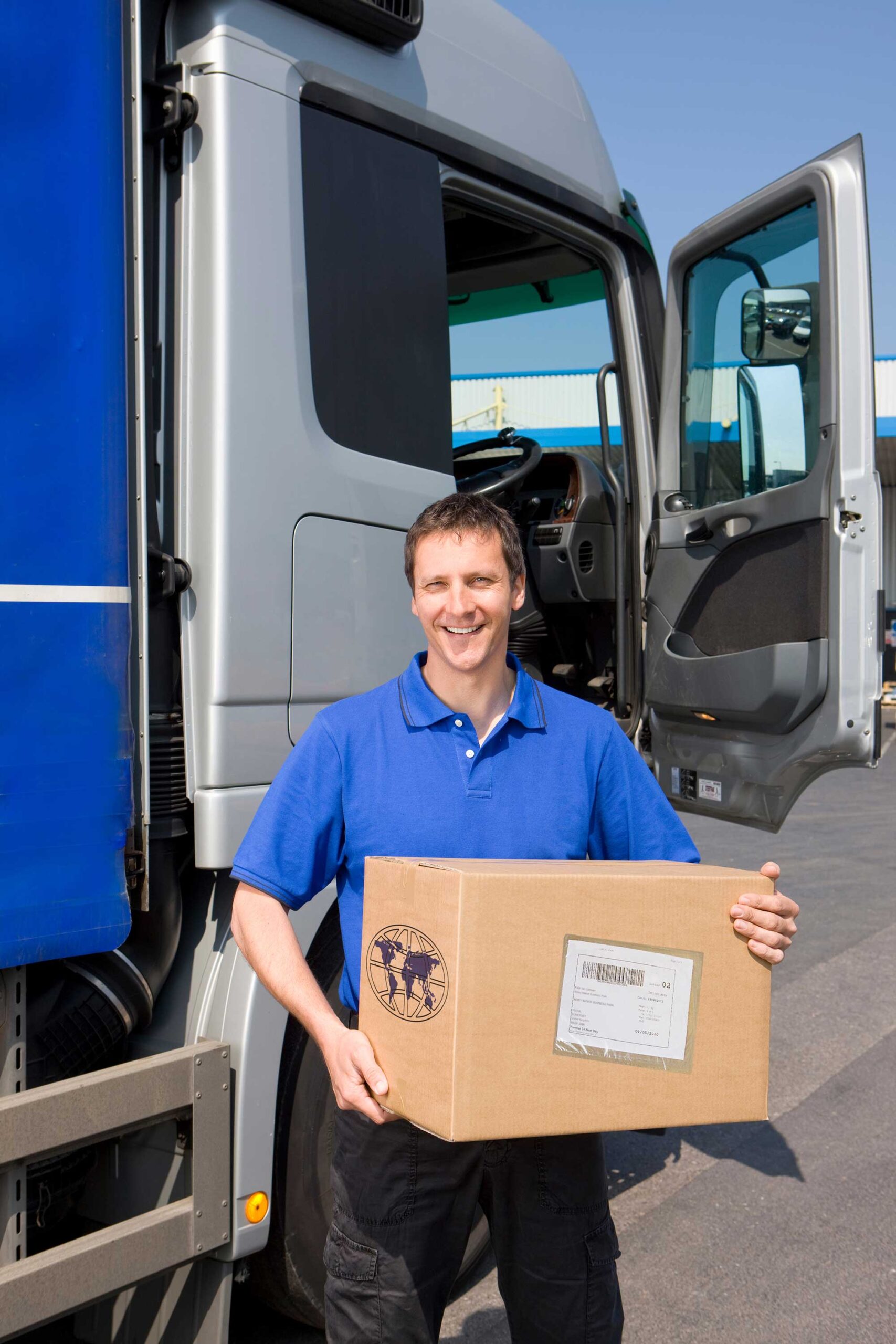 A man stands in front of a semi truck, holding a box with both hands